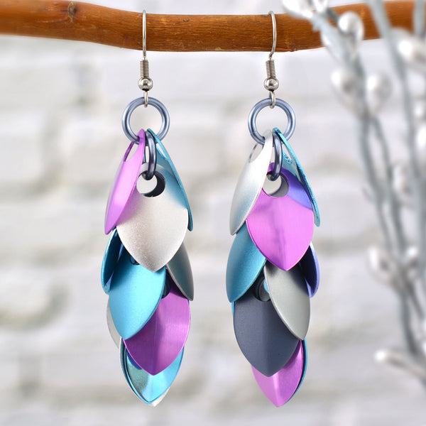 Long pastel metal leaf earrings by Rebeca Mojica on a branch. Each earring has 12 leaves (scales) hanging in pairs from a center row of 6 light grey links. Leaves are  silver, light blue, lilac and grey. The leaves stack in a feathered pattern