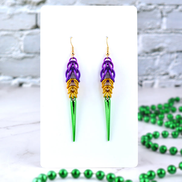 Chainmaille spike earrings in Mardi Gras colors hanging from a large white card. Earrings are on gold-plate earwire with purple at top (in gradually increasing link sizes), gold in the middle (tapering down in size), and a long shiny green spike at the base
