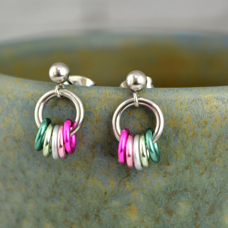 A pair of abrosexual pride chainmaille stud earrings balancing on the lip of a green marbled mug. Earrings have a steel link hanging from a tiny steel ball on the stud. On the steel link are 5 links in the colors of the abrosexual pride flag: shades of green, white and pink