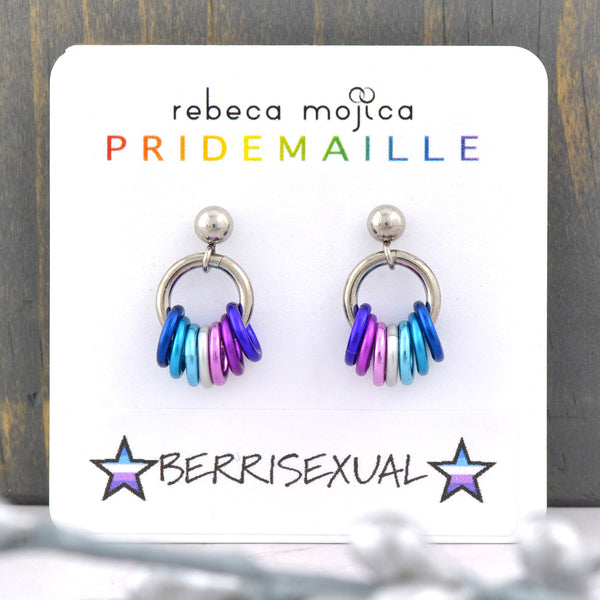 Tiny Berrisexual Pride chainmaille earrings displayed on a card. Earrings are on a post with a small steel ball. Fom the ball hangs a steel hoop that has 7 links hanging from it in the colors of the berrisexual pride flag: 3 shades of blue, white and 3 shades of purple/violet. The earring card reads Rebeca Mojica at top, the second line reads PRIDEMAILLE in rainbow color fonts. The bottom of the card has a sticker with the word BERRISEXUAL flanked by 2 stars in the color of the berrisexual flag