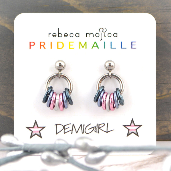 Demigirl earrings on a white card. These lgbtq pride stud earrings are made in colors to best match the demigirl flag - two shades of grey, white and pink. Displayed on a card by artisan Rebeca Mojica