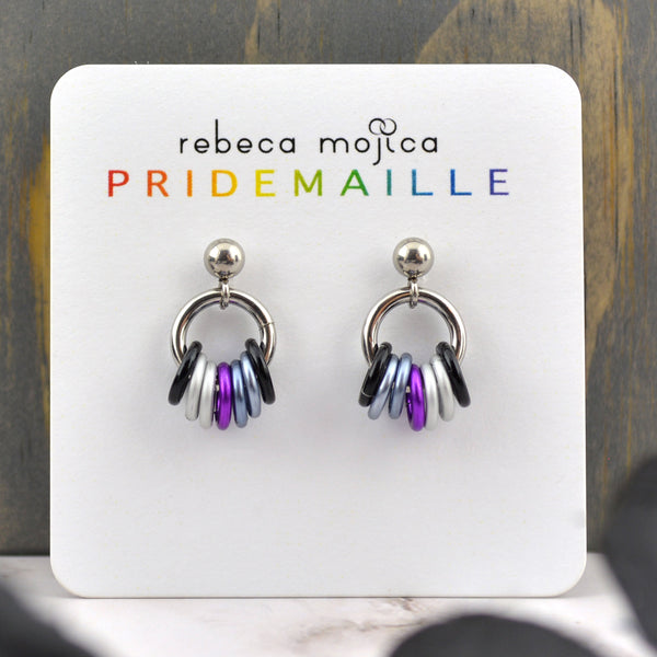 Tiny demisexual pride chainmaille earrings on a card. Earrings are on a small stainless steel stud with a ball. Hanging from the ball is a steel link with 7 smaller links in the colors of the demi pride flag (black, grey, violet and white). Card reads Rebeca Mojica PRIDEMAILLE