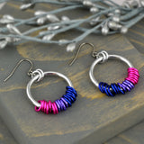 Bi pride earrings displayed on grey wood. The chainmaille earrings by Rebeca Mojica have a large silver color hoop, and on the silver hoop are 18 tiny links in the colors of the bisexual pride flag (7 links each of pink and blue, with 4 lilac links in between). The tiny colors are mirror image on the second earring.