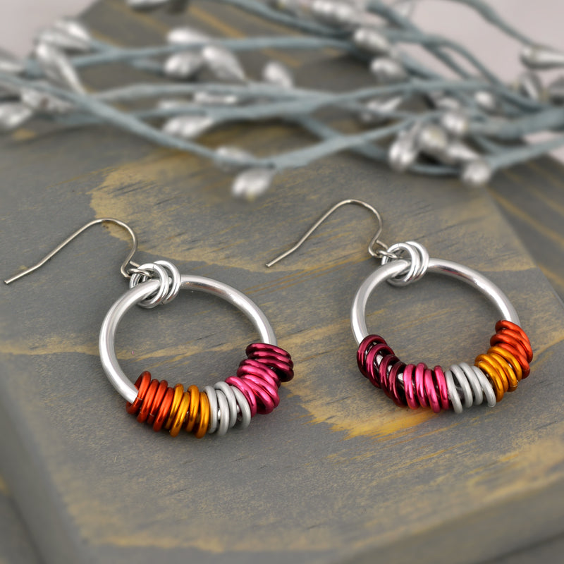 Lesbian pride earrings displayed on grey wood. The chainmaille earrings by Rebeca Mojica have a large silver color hoop, and on the silver hoop are 20 tiny links in the colors of the lesbian pride flag (4 links in each color: two shades of pink, two shades of orange + white). The tiny colors are mirror image on the second earring.