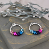 Omnisexual pride chainmaille earrings by Rebeca Mojica on grey wood. Earrings have a large silver color hoop, and on the silver hoop are 20 tiny links; four links in each of the 5 colors of the omnisexual pride flag (pinks, dark purple, blues).