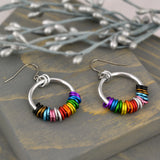 LGBTQ pride chainmaille hoop earrings by Rebeca Mojica on grey wood. Each earring has a large hoop link with 22 tiny links; two links in each of the 11 colors of the rainbow progress pride flag