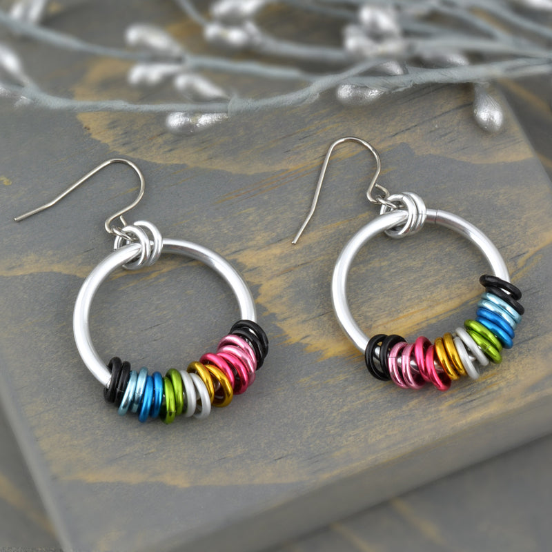 Closeup aerial view of chainmaille hoop earrings in colors of the queer pride flag. Earring has a large aluminum link with 18 hanging  tiny links, 2 links for each color of the queer pride flag: black, light blue, blue, lime, white, gold, dark pink, light pink, black