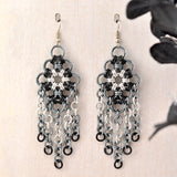 Closeup of chainmaille chandelier earring in silver, grey and black hanging from a wood backdrop. Medium and small links make a diamond shape, and hanging along the bottom of the the diamond are 5 strands of links in a silver to black ombre