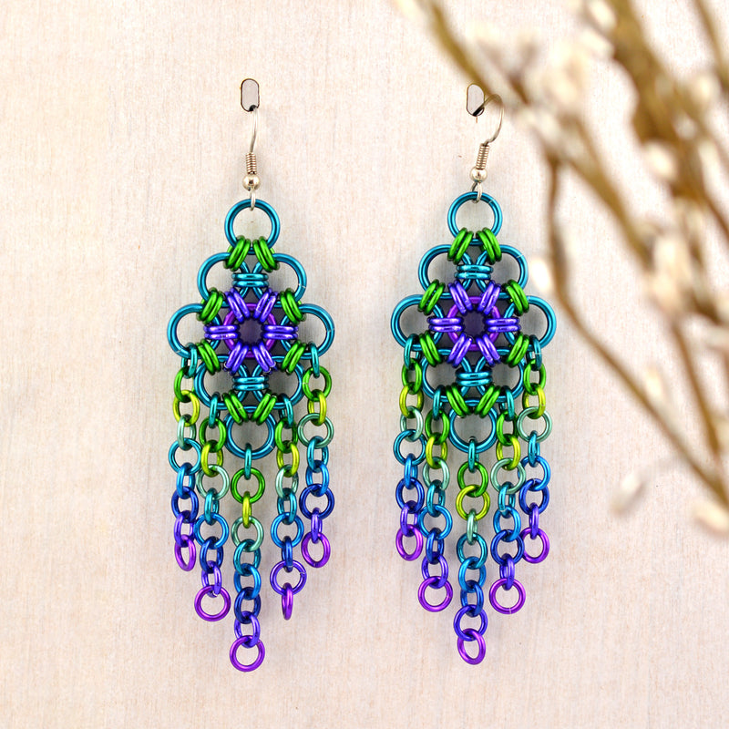 Long statement chainmaille earring in peacock colors on a light wood background. Earrings are shades of blue, green and purple in a chainmaille diamond shape with 5 strands of chainmaille links dangling beneath the diamond