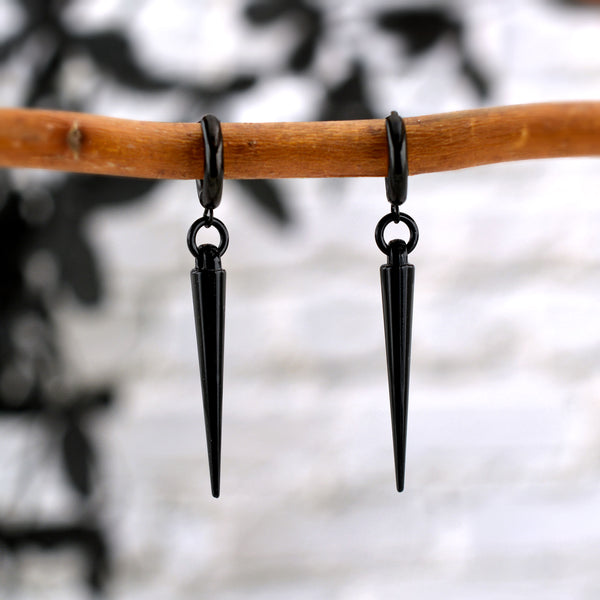 Pure black huggie earrings with black spikes. There is a small black ring connecting each huggie to a spike. These spike earrings by Rebeca Mojica are displayed on a branch with a mostly white and grey blurred background