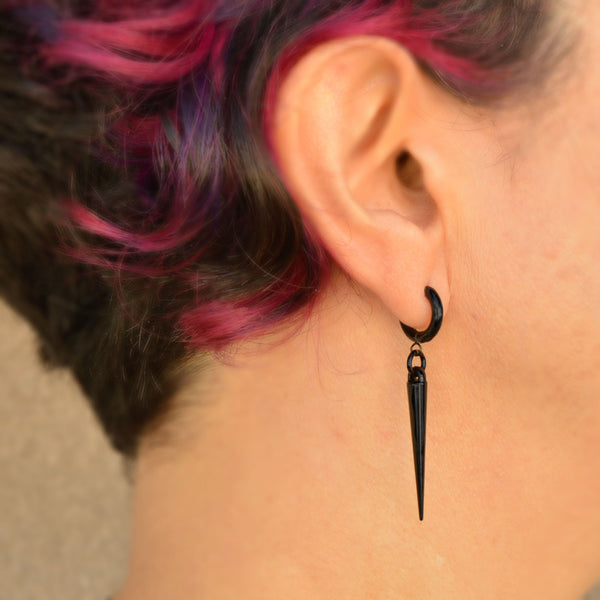 Profile/slightly rear angle view of a black spike hoop huggie being worn. There is a small black ring connecting the shiny black huggie to an acrylic spike. Artisan Rebeca Mojica is wearing the earring; her short red and black hair is visible at left