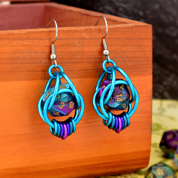 Chainmaille dice earrings by Rebeca Mojica featuring mini d20 in marbled purple and turquoise with gold numbers encased in a bright aqua aluminum cage. The bottom of the earrings have 7 tiny rings hanging side by side in a turquoise-purple-violet ombre. The earrings have stainless steel fishhook earwires and are hanging from a wooden box. 