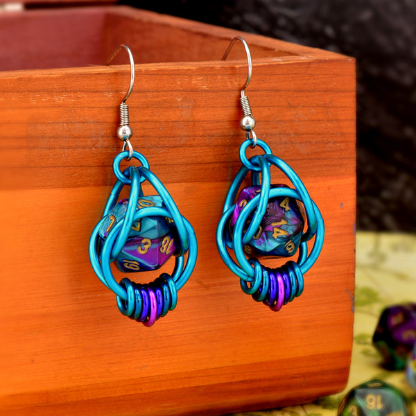 Chainmaille dice earrings by Rebeca Mojica featuring mini d20 in marbled purple and turquoise with gold numbers encased in a bright aqua aluminum cage. The bottom of the earrings have 7 tiny rings hanging side by side in a turquoise-purple-violet ombre. The earrings have stainless steel fishhook earwires and are hanging from a wooden box. 