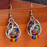 Closeup of chainmaille dice earrings by Rebeca Mojica hanging on a wooden box. Each earring has a mini multi-colored swirl 20-sided die with yellow numbers, encased in a 4-ring aluminum cage, with 6 tiny rings hanging from the bottom in the colors of the lgbtq rainbow pride flag. 