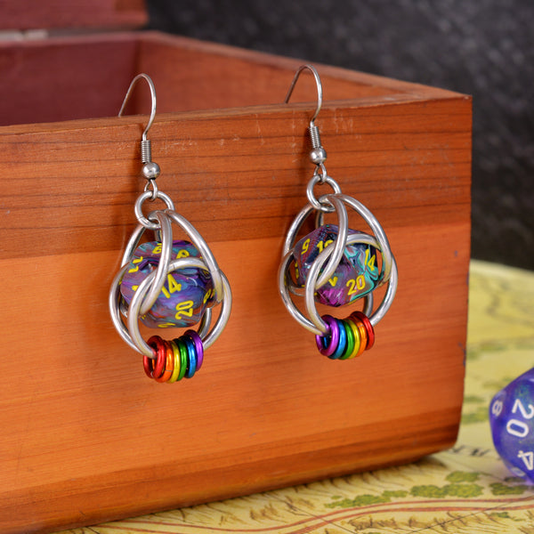 Closeup of chainmaille earrings by Rebeca Mojica hanging on a wooden box. Each earring has a mini multi-colored swirl 20-sided die with yellow numbers, encased in a 4-ring aluminum cage, with 6 tiny rings hanging from the bottom in the colors of the lgbtq rainbow pride flag. 