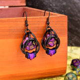 Chainmaille dice earrings by Rebeca Mojica featuring mini d20 in marbled purple and black with gold numbers encased in a black aluminum cage. The bottom of the earrings have 7 tiny rings hanging side by side in a black-purple-violet ombre. The earrings have black fishhook earwires and are hanging from a wooden box. 