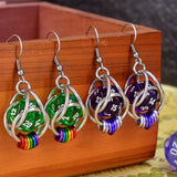 Closeup of chainmaille earrings by Rebeca Mojica hanging on a wood box. Each has a mini translucent 20-sided die in a cage of silver color aluminum links; the left pair has green dice with hanging rainbow links. The right pair has purple dice with purple ombre links.