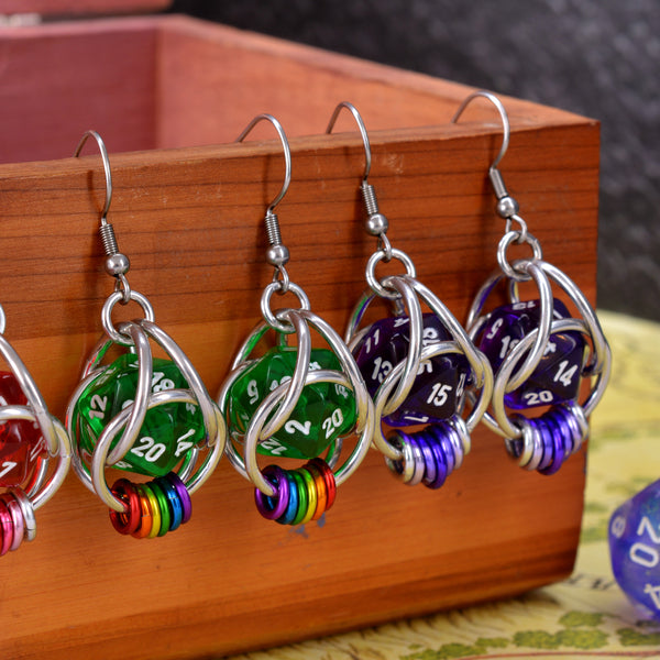 Closeup of chainmaille earrings by Rebeca Mojica hanging on a wood box. Each has a mini translucent 20-sided die in a cage of silver color aluminum links; the left pair has green dice with hanging rainbow links. The right pair has purple dice with purple ombre links.  A red earring is about 1/4 visible at far left.