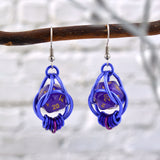 Chainmaille dice earrings by Rebeca Mojica hanging from a branch wood. Each earring has a mini purple 20-sided die with gold numbers, encased in a 4-ring purple aluminum cage, with 7 tiny rings hanging like a coil in shades of purple and violet