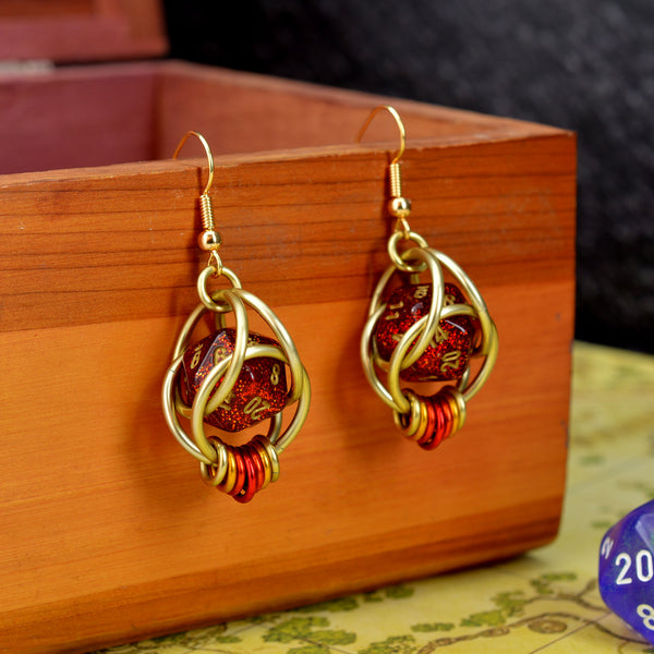 Closeup of chainmaille dice earrings by Rebeca Mojica hanging on a wooden box. Each earring has a mini red glitter 20-sided die with gold numbers, encased in a 4-ring light gold color aluminum cage, with 7 tiny rings hanging from the bottom in an ombre from gold to red and back to gold. 