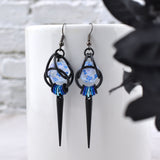 Chainmaille dice earring by Rebeca Mojica on a white mug. The earring has a translucent mini 20-sided die with blue numbers encaged in black aluminum links. At the bottom is an ombre of 7 blue to black links with a long black spike hanging from the lightest link in the center