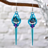 Chainmaille dice earrings by Rebeca Mojica featuring mini d20 in marbled purple and turquoise with gold numbers encased in a bright aqua aluminum cage. The bottom of the earrings have 7 tiny links hanging side by side in a turquoise-purple-violet ombre, with a bright turquoise spike hanging from the center link. The earrings have stainless steel fishhook earwires and are hanging from a branch against a light grey brick background.