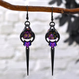 Chainmaille dice earrings by Rebeca Mojica featuring mini d20 in marbled purple and black with gold numbers encased in a black aluminum cage. The bottom of the earrings have 7 tiny rings hanging side by side in a black-purple-violet ombre, and a long black spike hangs from the middle link. The earrings have black fishhook earwires and are hanging from a branch against a light grey background