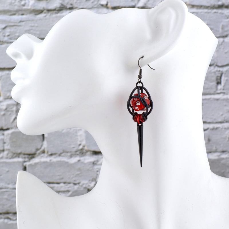 Chainmaille dice earring by Rebeca Mojica on a white display head. The earring has a translucent red mini 20-sided die with white numbers encaged in black aluminum links. At the bottom is an ombre of 7 red to black links with a long black spike hanging from the lightest red link in the center