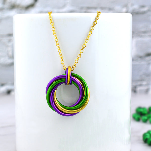 Chainmaille swirl pendant in mardi gras colors hanging from a white mug. Pendant has 6 large intertwined links—2 green, 2 purple and 2 gold—joined by a trio of smaller links: a small gold link flanked by 2 tiny purple links. The trio of links is attached to a thin gold chain. 