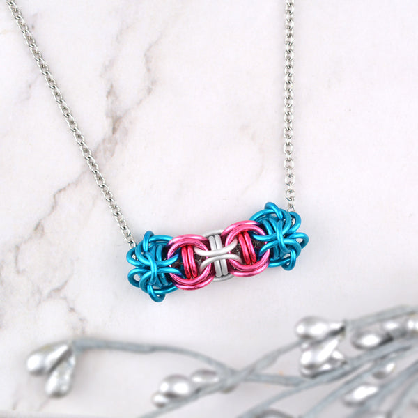 Transgender pride necklace displayed on a light marble surface. The chainmaille links form a small flexible tube (in the Captive Inverted Round pattern), about 1.5 inches long, that slides along the thin steel chain. Three links of a single color make a triangle that surrounds 2 of the same color link. There is one triangle for each stripe of the flag: blue, pink, white, pink and blue, and the triangles are joined to each other. Made by Rebeca Mojica in California, USA