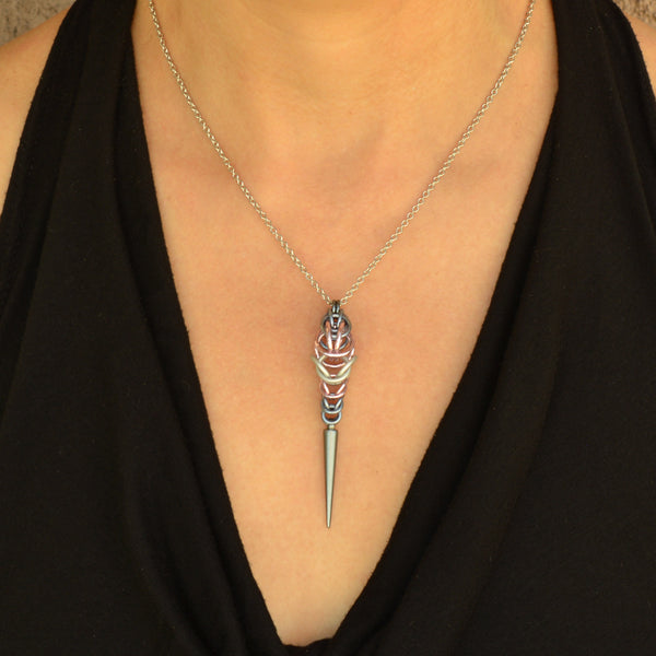 Demigirl pride pendant by Rebeca Mojica being worn. Pendant is chainmaille in the colors of the demigirl pride flag - grey, pink and white, with a light grey acrylic spike at the bottom.