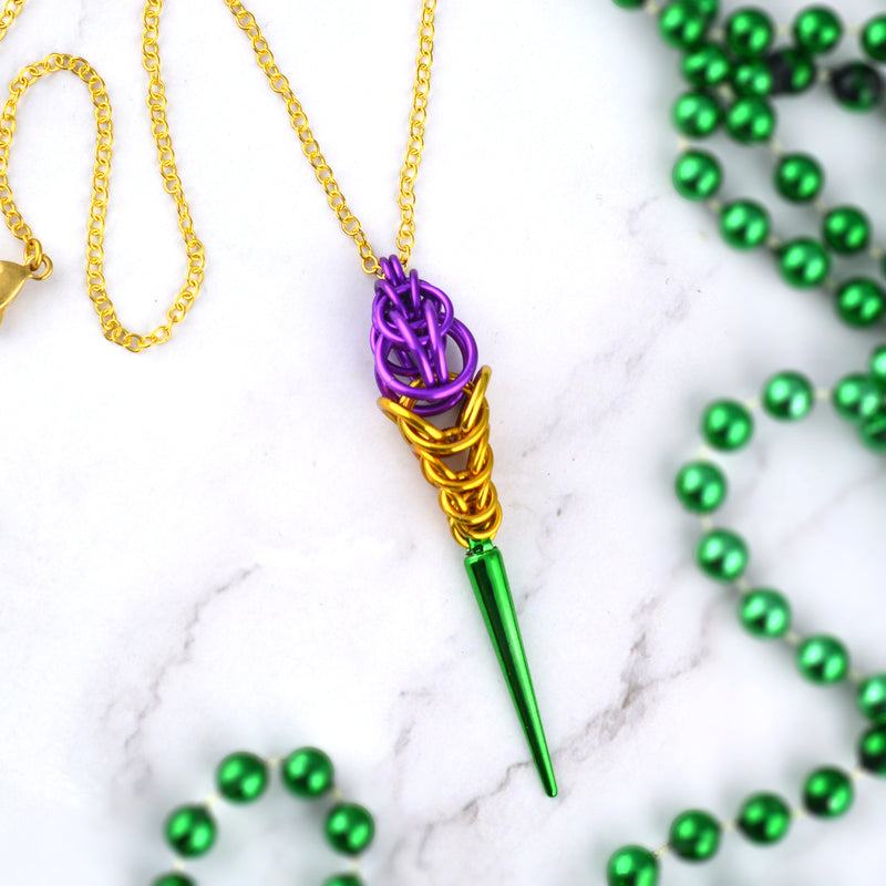 Aerial view of a Mardi Gras necklace on a white marble surface. Pendant has small chainmaille violet rings at top in increasing size, then gold rings of decreasing size, with a long green spike at the bottom. The Mardi Gras pendant is on a thin gold chain