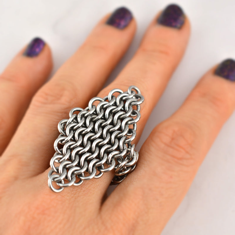 Closeup of a dense chainmaille stainless steel mesh ring being worn on a middle finger. The ring is diamond shaped and extends from below the finger to the first knuckle.