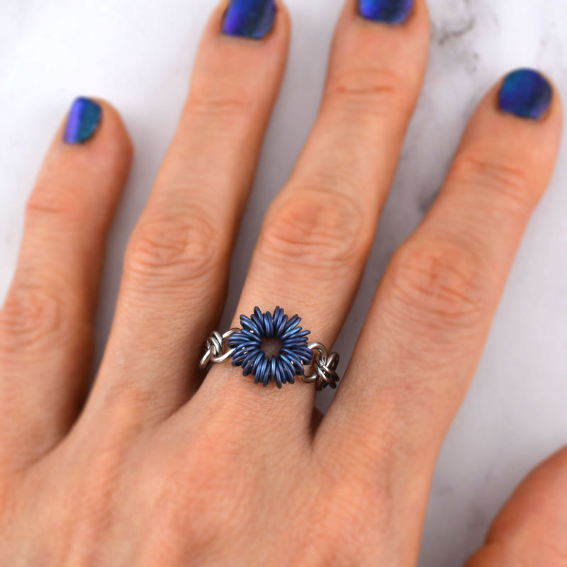 Closeup of blue titanium chainmaille ring by Rebeca Mojica being worn on the middle finger. Almost 40 blue links are arranged in a starburst pattern, in a circle around a larger, hidden steel ring. The band alternates medium sized steel rings with doubled tiny steel links.